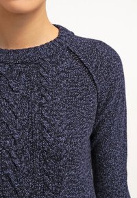 Mörkblå stickad tröja med en strukturerad kabelmönster, ribbat halsringning och raglanärmar. Tillverkad av ett mjukt, tjockt garn.