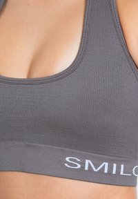 Sujetador deportivo gris con textura acanalada, que cuenta con una banda elástica ancha con "SMILO" grabado en blanco. El diseño tiene un escote redondeado y tirantes anchos.