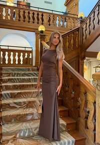 Femme dans une robe marron ajustée tenant une pochette beige, debout sur un escalier en bois moquetté avec des rampes ornées et un éclairage chaleureux.