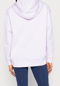 Person som bär en enkel ljuslila hoodie och mörkblå leggings, visad bakifrån med händerna avslappnat vid sidorna.