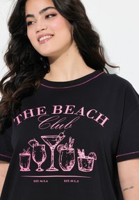 Camiseta negra con costuras rosas, que presenta una impresión gráfica de cócteles y el texto "THE BEACH Club" en letras rosas en negrita.