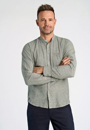 Camisa verde de botonadura con cuello mao, bolsillo en el pecho y tejido texturizado, combinada con pantalones oscuros. El modelo está de pie con los brazos cruzados.