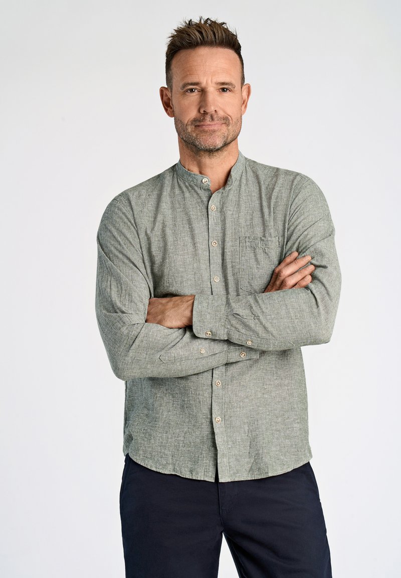 Camisa verde de botonadura con cuello mao, bolsillo en el pecho y tejido texturizado, combinada con pantalones oscuros. El modelo está de pie con los brazos cruzados.