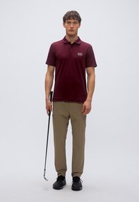 Πόλο πουκάμισο μπορντό με κοντά μανίκια και λογότυπο, συνδυασμένο με μπεζ παντελόνια με πλαϊνές τσέπες. Το μοντέλο κρατάει ένα γκολφ κλαμπ και φοράει μαύρα παπούτσια.