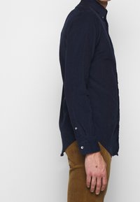 Muže oblečeného v námořnicky modré košili s dlouhými rukávy a hnědých kalhotách, z profilu, na jednobarevném světlém pozadí.