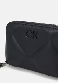 Black quilted leather wallet with embossed "CK" logo, featuring a silver zipper and textured surface pattern. Compact, rectangular shape.