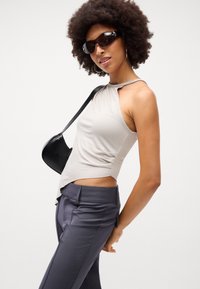 Top senza maniche beige con orlo asimmetrico, abbinato a pantaloni grigi su misura. Una borsa a tracolla nera e occhiali da sole scuri completano il look.
