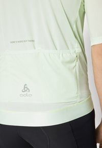 Jasnozielona koszulka sportowa z teksturowanym wzorem, z logo na dole i kieszenią na zamek. Zawiera motywacyjny nadruk na plecach.