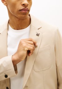 Άνδρας ρυθμίζει το καρφίτσα στο πέτο μπεζ σακακιού, φορά λευκό πουκάμισο με στρογγυλή λαιμόκοψη, με μπεζ σακάκι που έχει τσέπη στο στήθος και μανσέτα με μαύρα κουμπιά.