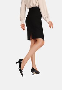 Gonna nera a matita con spacco posteriore, orlo in pizzo texturizzato, abbinata a scarpe nere lucide con tacco medio. Tessuto liscio e design attillato.