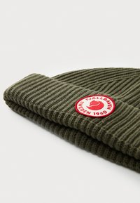 Grön stickad mössa med ribbad textur, som har en vikbar kant och en cirkulär röd logotyp med vit text.