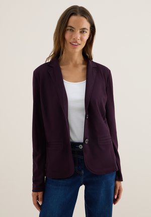 Blazer violet en tissu extensible, avec une fermeture à un bouton, des revers en pointe et deux poches avant. Porté sur un haut blanc.