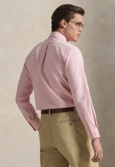 Man met een bril, een lichtroze shirt met lange mouwen en beige broek, van achteren gezien, draait zijn hoofd opzij.