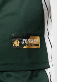 Groene sportkledingstof met een zwart en gouden label met een gorillagrafiek en de tekst: "TEAM GORILLA WEAR VOOR DE GEMOTIVEERDEN SINDS 1982."