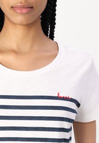 Žena v bílém tričku s námořnickými modrými vodorovnými pruhy a malým červeným vyšitým slovem "love" poblíž límce na levé straně.