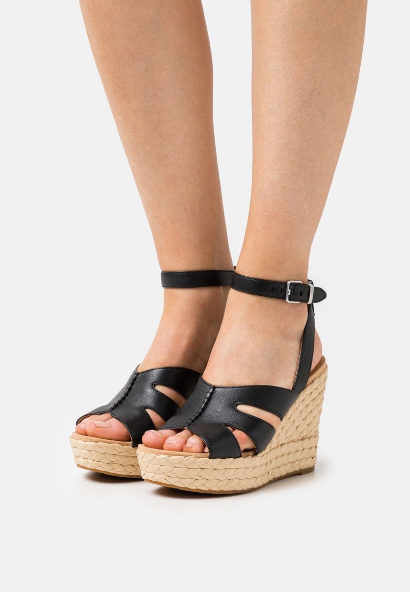 Jambes portant des sandales compensées à bride cheville en cuir noir avec des semelles plateforme en espadrille tissée sur un fond blanc.