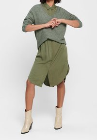 Nainen, joka on pukeutunut oliivinvihreään paitamekkoon harmaan villatakin ja beigeihin korkokenkiin, seisoo kädet ristissä ja jalat eristyksissä.