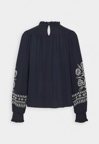Navyblaue Bluse mit langen Ärmeln, die über aufwendige weiße Stickereien an den Ärmeln sowie einen gerafften Ausschnitt mit einem Schlüsselaugenverschluss auf der Rückseite verfügt.
