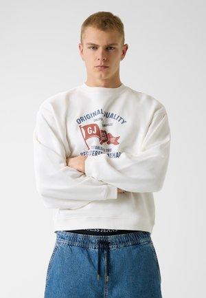 Junger Mann mit kurzen blonden Haaren und Ohrringen, der ein weißes Sweatshirt mit rotem und blauem Logo trägt und die Arme verschränkt stehen.