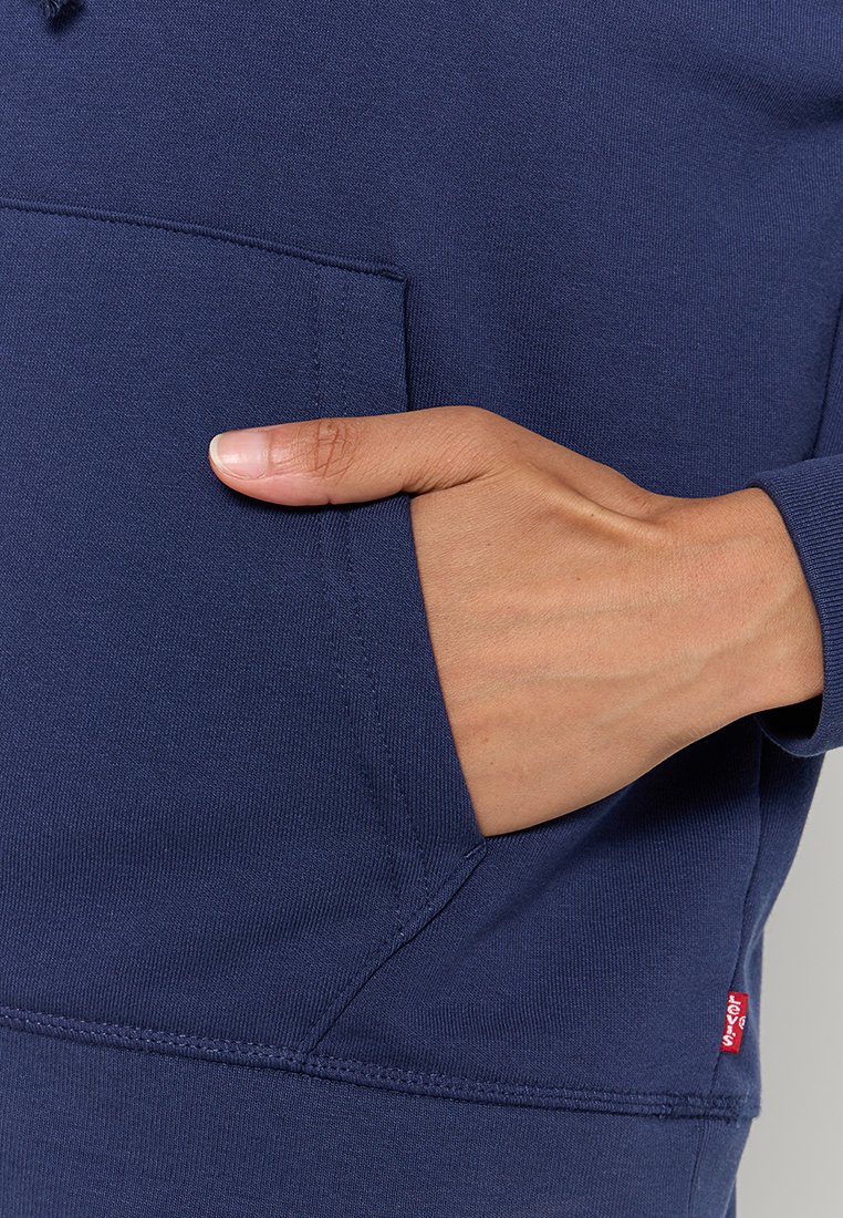 Sudadera azul marino con un bolsillo frontal, puños acanalados y etiqueta de logo en el dobladillo. Tejido suave con una textura lisa, diseño ajustado.