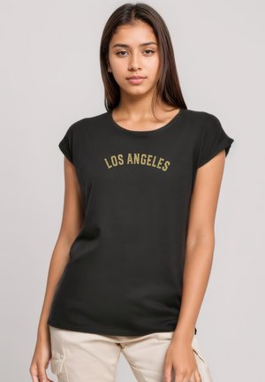 Young woman standing, wearing a black t-shirt with "LOS ANGELES" text and light beige cargo pants against a plain background.