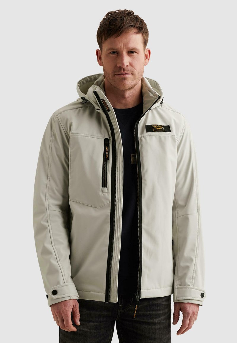 Mann trägt hellgraue Kapuzenjacke mit Reißverschluss über schwarzem Hemd, steht vor einfarbigem hellem Hintergrund.