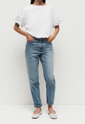 Osoba nosząca białą bluzkę z krótkim rękawem wpuszczoną w niebieskie dżinsy z wysokim stanem, ozdobione perełkami, oraz białe plecione płaskie buty.