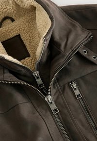 Braune Lederjacke mit einem hellen Fleecefutter, ausgestattet mit silbernen Reißverschlüssen und einem hohen Kragen, aus einer Draufsicht präsentiert.