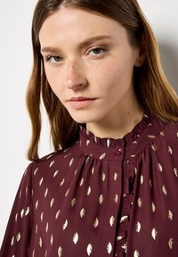 Burgundy blouse featuring a ruffled collar, button closure, and metallic gold leaf patterns across the fabric. Smooth texture.