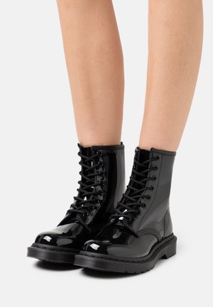 Bottines à lacets en cuir verni noir brillant portées sur des jambes nues sur fond blanc uni.