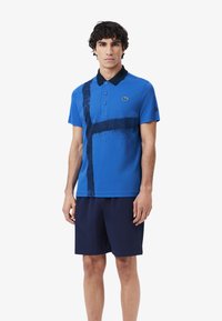 Blå polotröja med ett svart abstrakt mönster, tre knappar och en kontrasterande mörk krage. Bärs med mörka shorts.