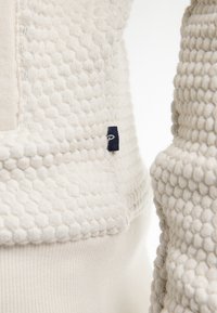 Pohodlný béžový svetr s texturovaným vzorem bobble-pletení, který má hladkou pletenou část a malou námořnickou štítkovou značku na boku.