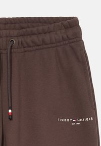 Primer plano de pantalones de chándal marrones con cinturilla elástica, cordón, bolsillo lateral y pequeño logotipo de Tommy Hilfiger con los colores de la marca y "Est. 1985."