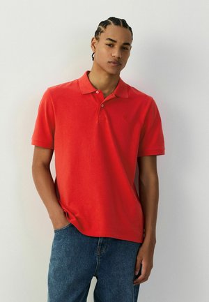 Junger Mann mit geflochtenem Haar, der ein rotes Poloshirt mit kurzen Ärmeln und blaue Jeans trägt, vor einem schlichten weißen Hintergrund stehend.