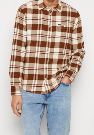 Person trägt braun-weiß kariertes Hemd mit Brusttasche und hellblaue Jeans, Hand in der Tasche.