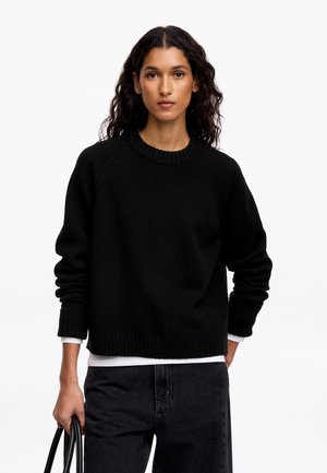 Femme aux cheveux bouclés foncés porte un pull noir sur une chemise blanche et un jean noir, tenant un sac à main noir sur fond blanc.