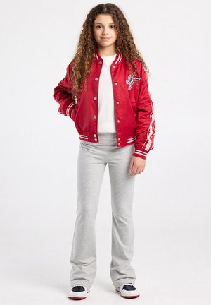 Jonge persoon met krullend haar, gekleed in een rood jasje, wit overhemd, lichtgrijze broek en witte sneakers, staand met één hand in de zak.