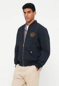 Tumesinise värvi pommikindel jakk, millel on kuldsete tikanditega logo, tõmblukk, küljetaskud ja soonikkoes mansetid, kantud kontrollitud särgi üle.