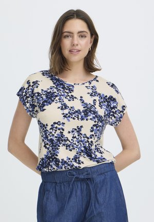 Młoda kobieta ubrana w beżową bluzkę z krótkim rękawem w niebieskie kwiatowe wzory oraz niebieskie, wysokie spodnie z wiązaniem, stojąca na jednolitym tle.