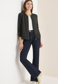 Ciemnozielona kurtka bomberka z elastycznym dołem, biała koszulka, ciemnoniebieskie dzwony z kontrastowym przeszyciem, kremowe sneakersy. Proste tło.