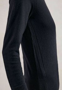 Primo piano di una persona che indossa un maglione aderente nero in maglia, con dettagli sulla cucitura laterale visibili e polsini e orlo a costine.
