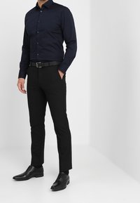Hombre de pie con una mano en el bolsillo, vistiendo una camisa ajustada de botón color marino, pantalones negros, cinturón negro y zapatos de vestir negros.