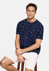 Hombre sentado en un taburete de madera, vistiendo una camiseta azul marino con un patrón de peces claros y pantalones cortos blancos contra un fondo liso.