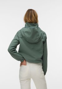 Frau mit schulterlangem Haar, die eine grüne Kapuzenjacke und weiße Hose trägt, steht mit einer Hand in der Gesäßtasche.