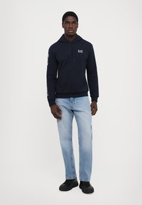 Felpa blu navy con tasca a marsupio frontale e cappuccio con cordino, abbinata a jeans azzurri e calzature nere.