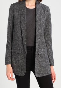 Texturerad grå blazer med sjalkrage, två framfickor och en skräddarsydd passform, bärs över en mörk skjorta och svarta byxor.