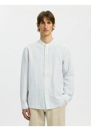 Ung mand med mellem langt brunt hår iført en lys, lodret stribet skjorte med knapper og beige bukser, der står mod en hvid baggrund.