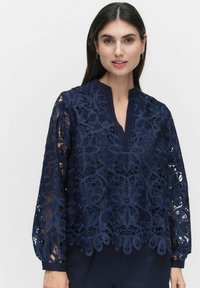 Granatowa koronkowa bluzka z kwiatowymi wzorami, dekoltem w kształcie litery V i długimi rękawami. Materiał jest półprzezroczysty, ukazujący skomplikowane wzory.