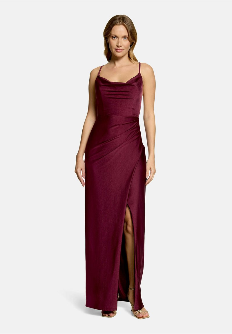 Femme portant une longue robe bordeaux sans manches avec une fente haute à la cuisse et des bretelles fines, se tenant devant un fond uni.
