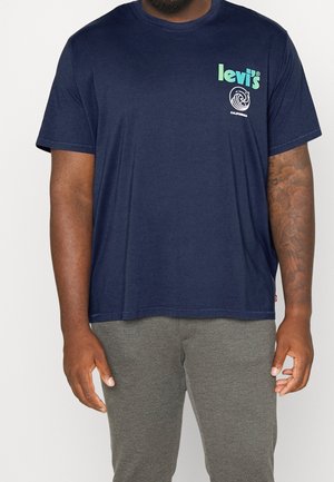 Homme portant un t-shirt Levi's bleu marine avec un logo vert et un motif de vague, associé à un pantalon gris, montrant un bras droit tatoué.